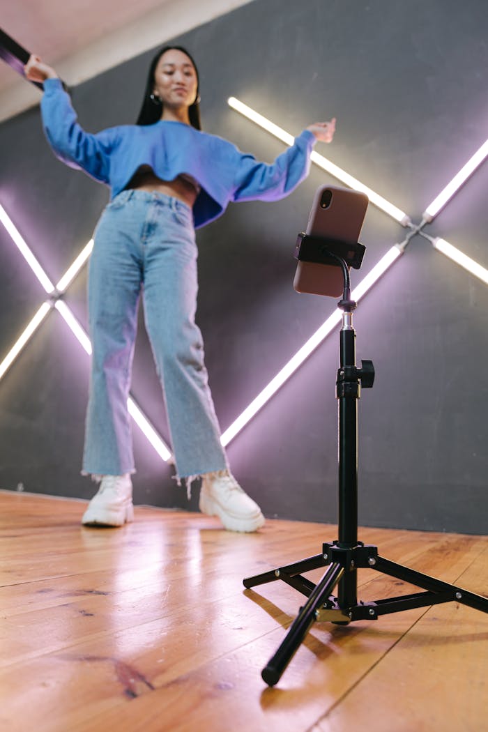 A young woman dances energetically in front of a smartphone camera on a tripod indoors.